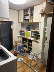 キッチンの残置物撤去前　棚に詰められた小物があふれ、床に踏み台、モップなどが散乱している