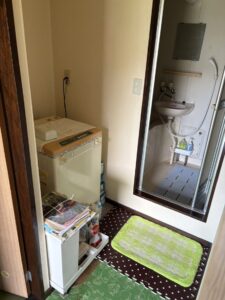 片付け前のバスルーム。脱衣所には洗濯機と棚が置かれており、ユニットバスには洗剤とマットが残されている。