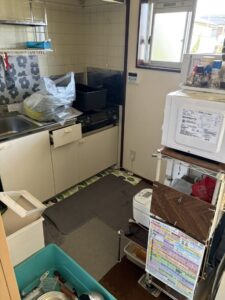 片付け前のキッチン。シンク回りに物が置かれ、収納ラックにもキッチン家電や生活用品が残されている。
