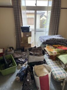 片付け前の寝室。布団が積まれたベッドの横に衣類やダンボールが置かれている。