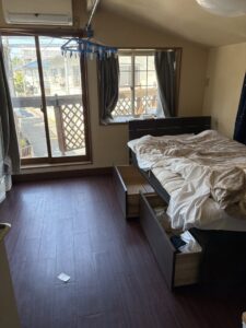 作業前の寝室。引出付ベッドの上に布団がかかっている。窓にはカーテンが残されカーテンレールから洗濯ハンガーがぶら下がっている
