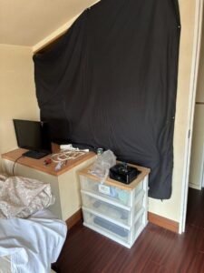 作業前の寝室。ベッドの足元に小型テレビや生活用品が入ったプラケースが置かれており、壁に黒くて大きな布がかかっている。