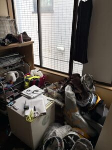 作業前の室内。1ドア冷蔵庫の上に文具が置かれ床には布製品をはじめとする生活用品が積まれている。